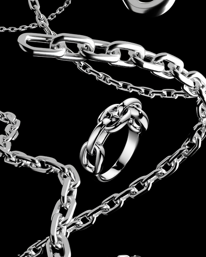 A close-up studio shot of a silver-toned chain necklace with chunky interlocking links, a matching ring, and a small oval charm, arranged on a black background.
