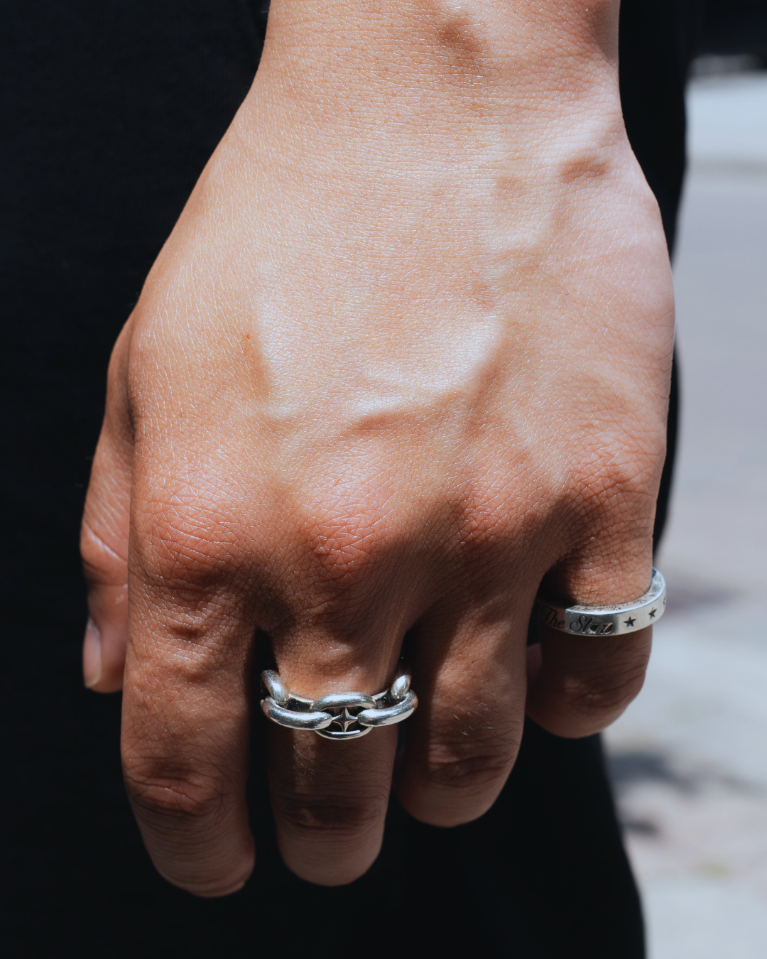 A close-up of a person's hand wearing two silver rings, one a textured band and the other a chain-link style ring, set against a dark black garment background.